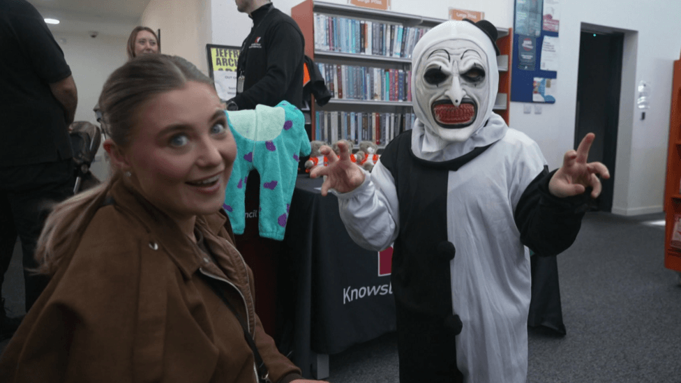 Woman with light brown hair tied in a ponytail, wearing a brown jacket, looks through a rail of donated Halloween costumes at Prescot Library. Other adults and children are gathered in the library, with bookshelves and a display of comics visible behind them.