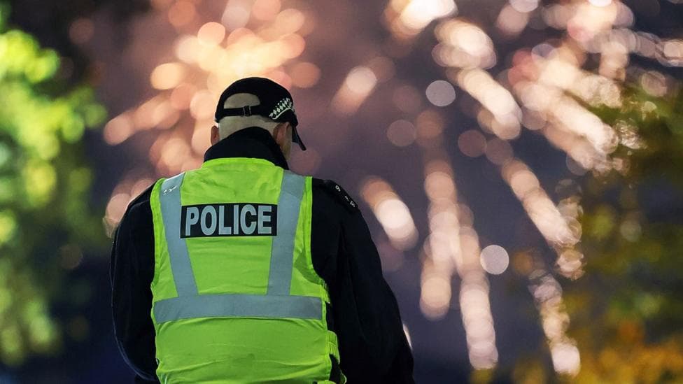 A male police officer wearing a black cap and a hi-vis jacket with his back to the camera. In the background, fireworks explode brightly against a dark night sky.