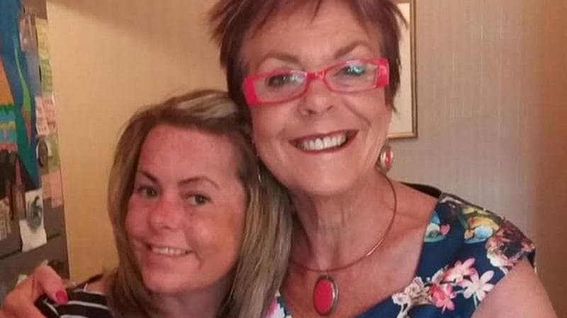 Clare and her mum standing indoors and embracing for a photo. Clare wears a navy-and-white striped top, while Paula wears a blue floral dress with bright pink glasses and matching jewelry
