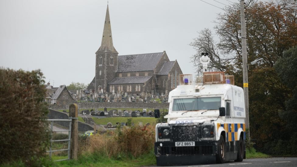 A PSNI landrover is parked on a rural road. It is white with blue and yellow checks. A church is in the background.