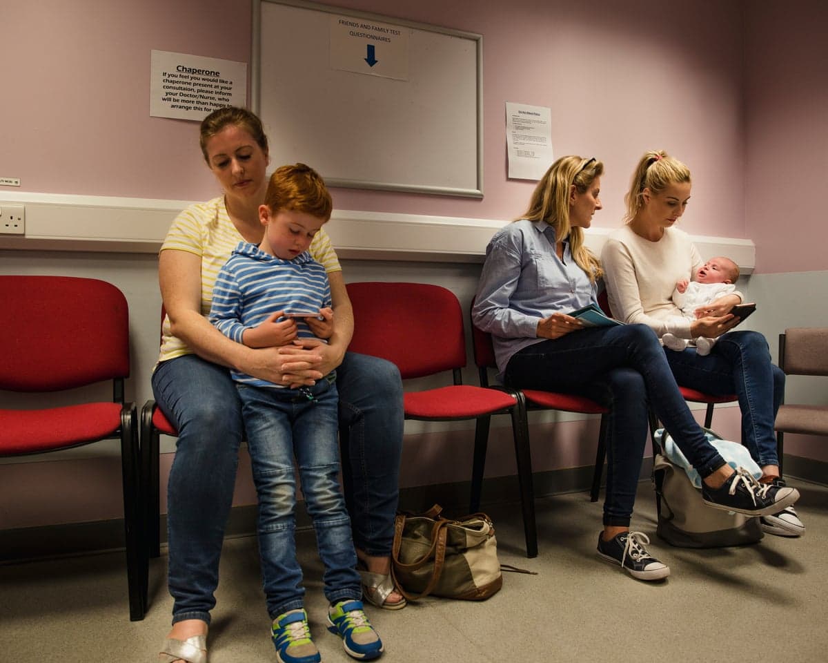A mother with a child sits next to another family with children in a row of red chairs in a doctor's waiting room