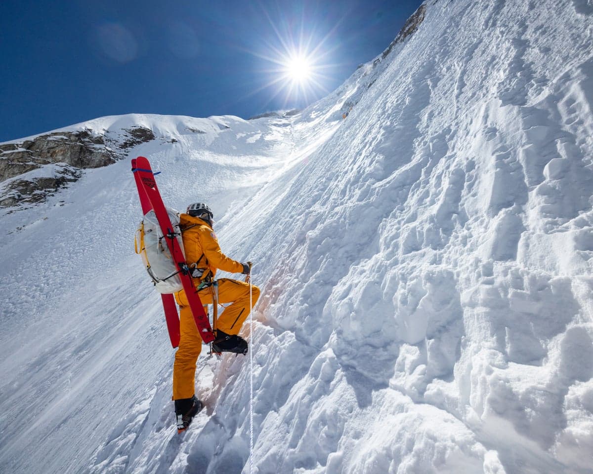 Jim Morrison carries red skis while climbing a steep snowy slope on Everest