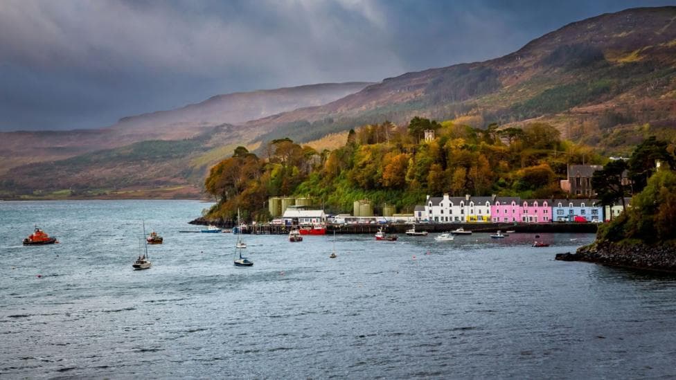 A view of Portree across the sea. There is a row of colorful, three-storey buildings along the harbour and boats, including an orange lifeboat, dot the harbour and the wider bay. Tree covered hills rise from behind the houses.