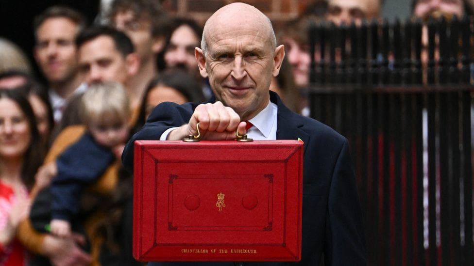 John Healey, who is bald, holds up a red ministerial box in front of a crowd on Downing Street