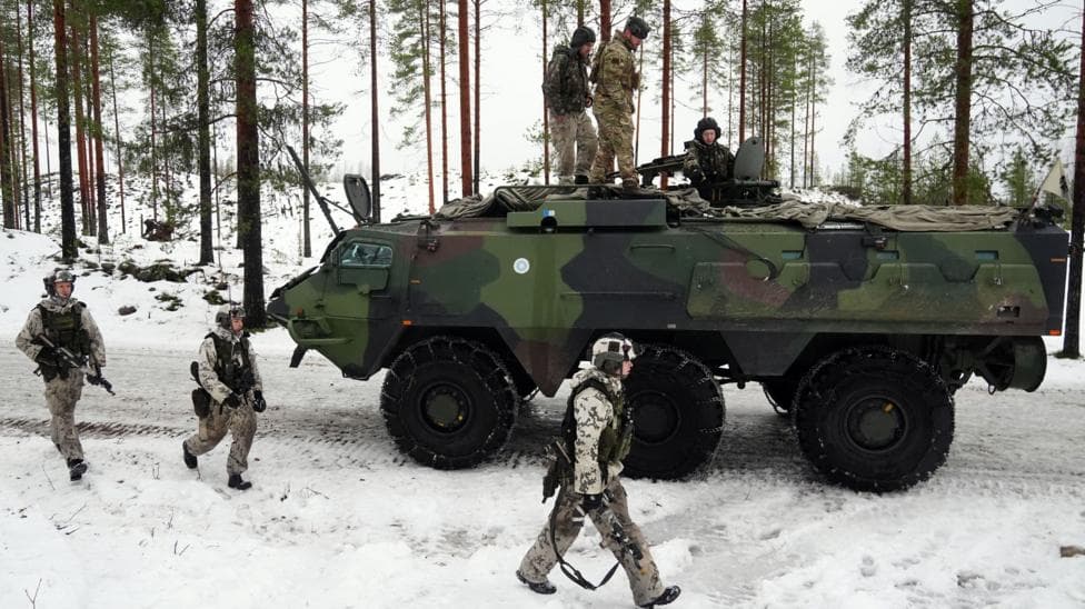 Three soldiers walk alongside an armoured tank in a snowy landscape. Three soldiers sit on top of the tank.