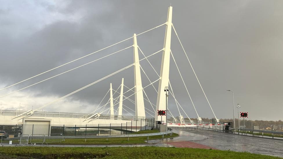 A river bridge, with a barrier down on one side to stop traffic going through at that time,The bridge is coloured white, with a grey overcast sky above it.
