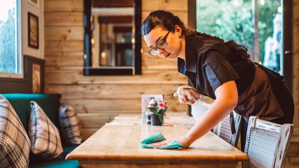 A young women working in a cafe wipes down a wooden table with a green cloth