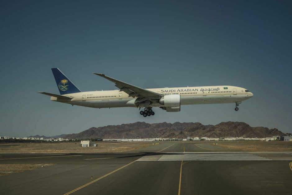 A Saudi Arabian Airlines Boeing 777 landing on a runway with mountainous backdrop.