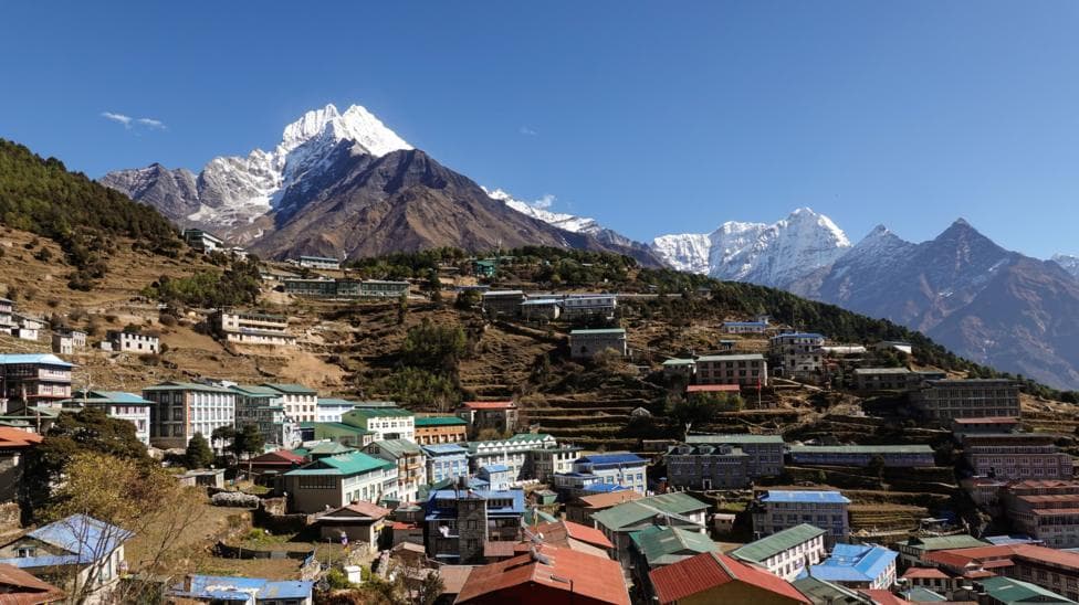 Namche Bazaar's colourful rooftops spread across terraced hillsides in Nepal's Everest region, with the snow-capped peak of Mount Thamserku towering above the town under a clear blue sky.