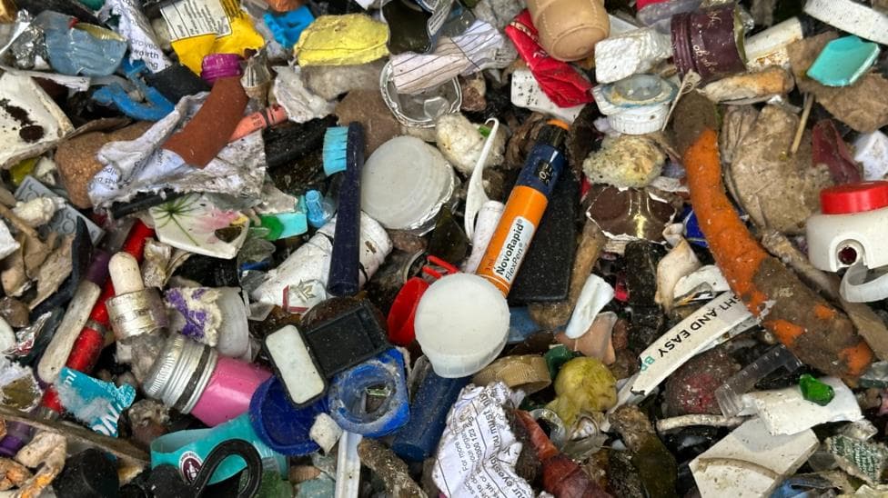 Close-up of mixed waste showing items including plastic bottle caps, containers, fragments, broken ceramics, pieces of glass, paper, and other household refuse. Various colours and textures are visible throughout the pile, including blue, red, yellow, white and green plastics. A carrot and other food waste can also be seen among the materials.