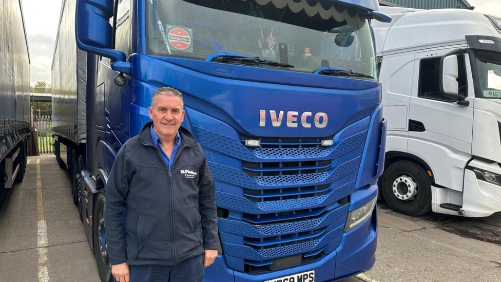 Martin Pinches is stood in front of a blue lorry. He is wearing a blue polo shirt and a navy jacket. He has short grey hair.
