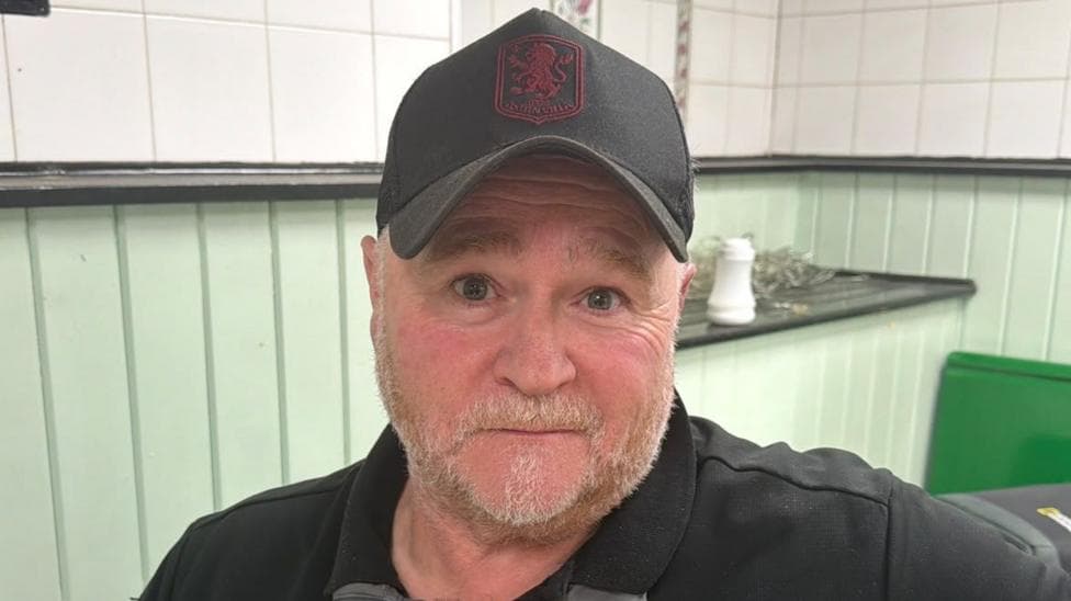 A man in a black cap, black zip hoody is looking directly at the camera