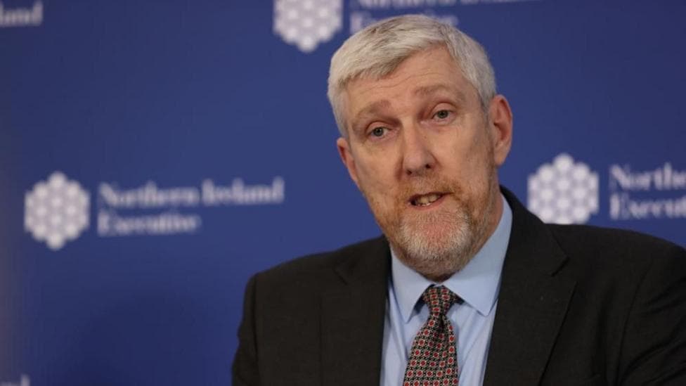 A man with short white hair and a white and ginger beard is standing in front of a blue background. The background is faded. He is wearing a dark suit, red and green patterned tie and a light blue shirt.