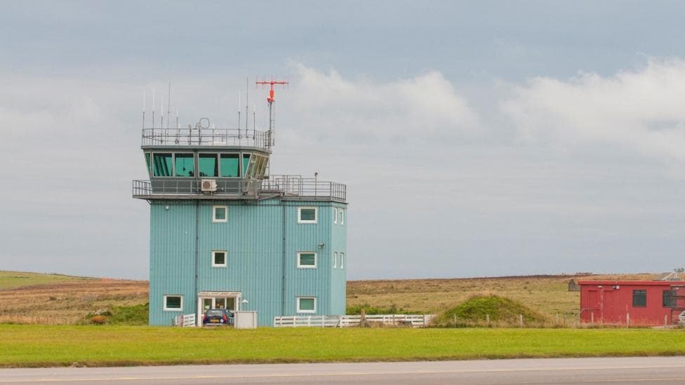 Control tower at Kirkwall Airport. The building is a pale blue-green colour with a control with large windows at the top. The tower is in a flat, grassy landscape.