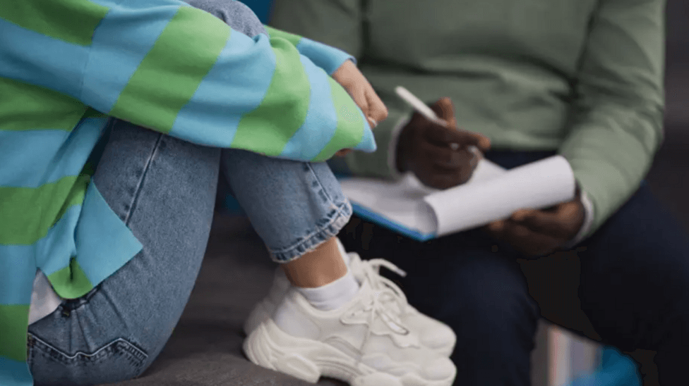 A person on the left is wearing jeans, a blue and green striped top, and white trainers. They are sitting on a chair with their legs pulled up. To their right is a person with a green top and navy trousers sitting in a chair with a clipboard and a pen. Neither person's head and shoulders is visible.