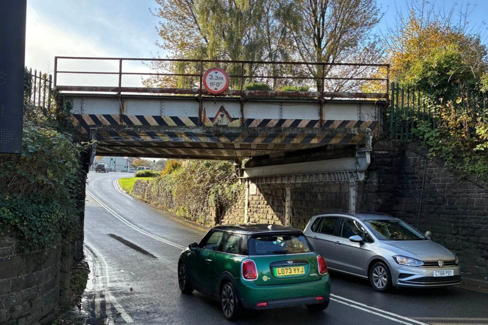 A road passes beneath a low railway bridge on a bright, sunny day. Two cars are travelling under the bridge, which is made of steel and stone and spans the road overhead. A circular height-restriction sign fixed to the bridge warns that the clearance is 3.3 metres (11 feet). The road surface appears wet and reflects the sunlight. Trees with autumn foliage rise above the bridge against a clear blue sky, while stone retaining walls and vegetation line both sides of the road.