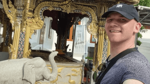 Rory McColl , who is wearing a black baseball cap, takes a selfie in front of a stone elephant and a historic building