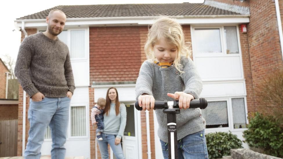 Family outside a house including a man, woman with a small child on her hip and another small child riding a scooter in the foreground