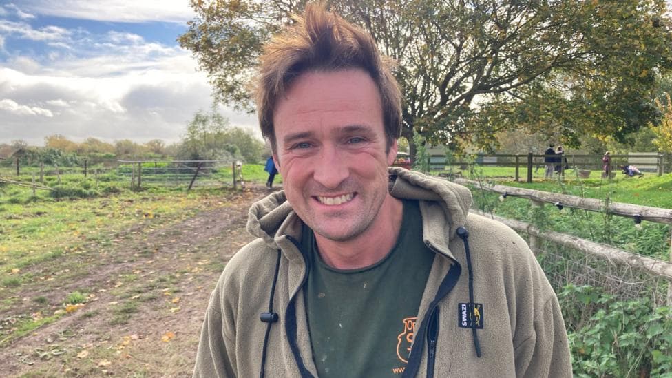 A man with short brown hair, a grey fleece and a green t-shirt smiles as he stands in a farm. A fence and a tree can be seen behind him.