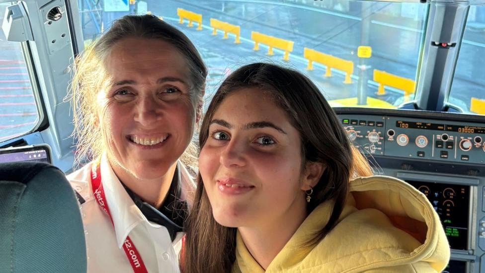 A woman wearing a white shirt and a Jet2 lanyard and a young woman wearing a yellow hooded top smile next to each other while in a plane's cockpit.