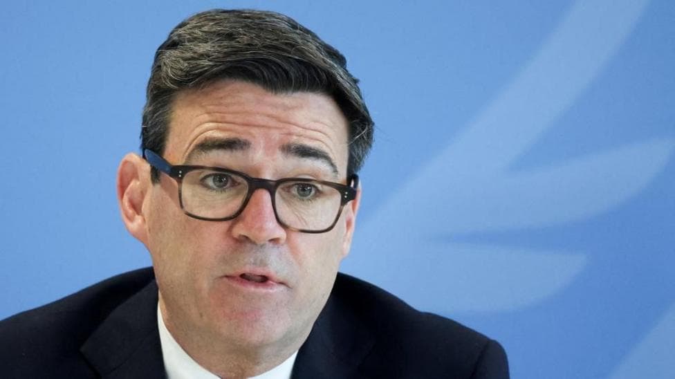 Andy Burnham is wearing a white shirt, dark-coloured jacket and glasses. He has short black hair and is sat in front of a blue backdrop.