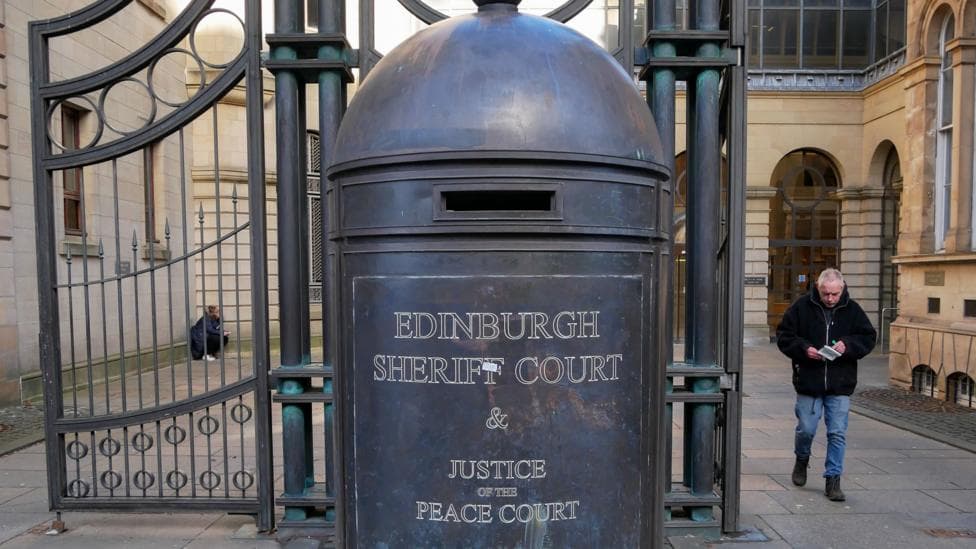 An older styler postbox which is dark in colour and has a letterbox, that has the words "Edinburgh Sheriff Court and Justice of the Peace Court". This is central to the image, and there is a gate on the left, and a man leaving the court on the right.