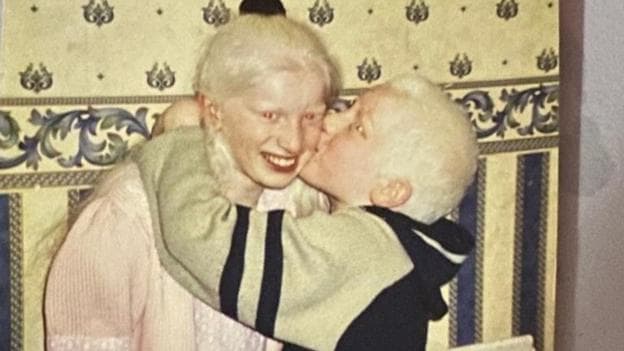 An old photo of Musarat Begum and Mohammed Rafi. Musurat is smiling and has long white hair tied in a ponytail, and a pink and white dress. Her brother has his arms around her neck and is kissing her cheek. He wears a beige and black hoodie and has white hair