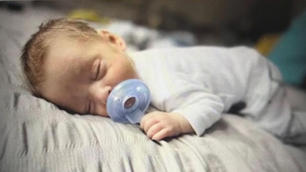 Baby lying on his front wearing a white a yellow striped baby gro with a circular plastic item infront of his mouth.