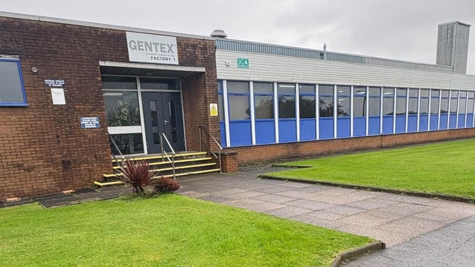 A single-storey flat-roofed building. There is a sign saying Gentex over the door, and a long line of windows to the right. There is a well-tended lawn to the front.