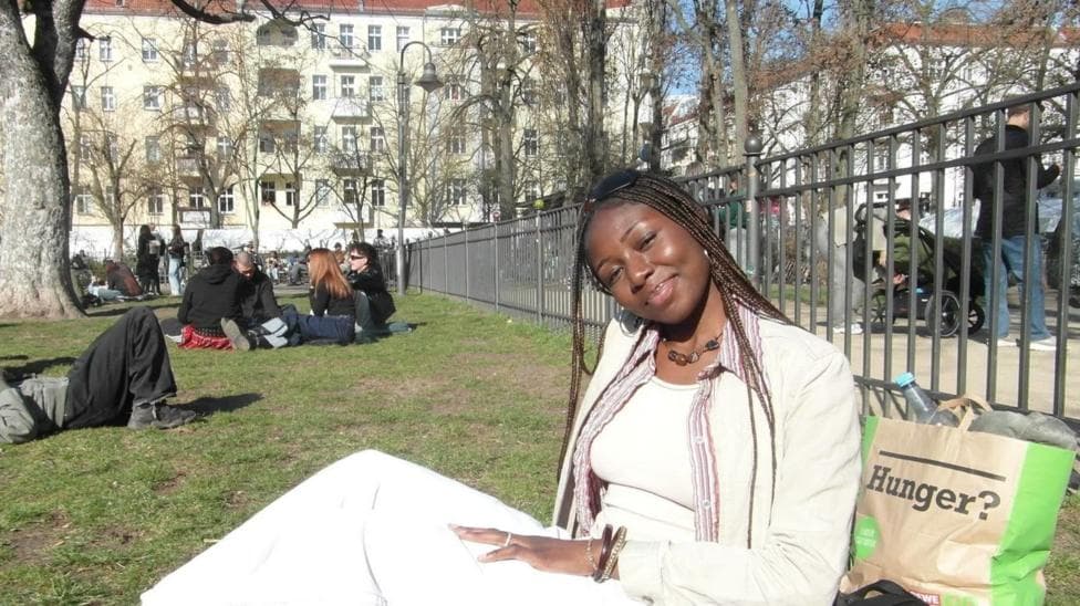 A young woman with long braids is sitting on the grass in a park on a sunny day. She is tilted slightly to her left, smiling gently toward the camera. She is wearing a cream top, jacket and trousers. Behind her is a paper bag with the word Hunger on it. In the background is a group of young people sitting on the grass
