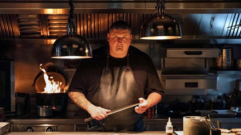 Gareth Ward in black chef overalls holding large knife with kitchen appliances and open fire behind him and large overhead light pendants. He has a tattoo down one arm and wears a leather apron and has short dark hair.