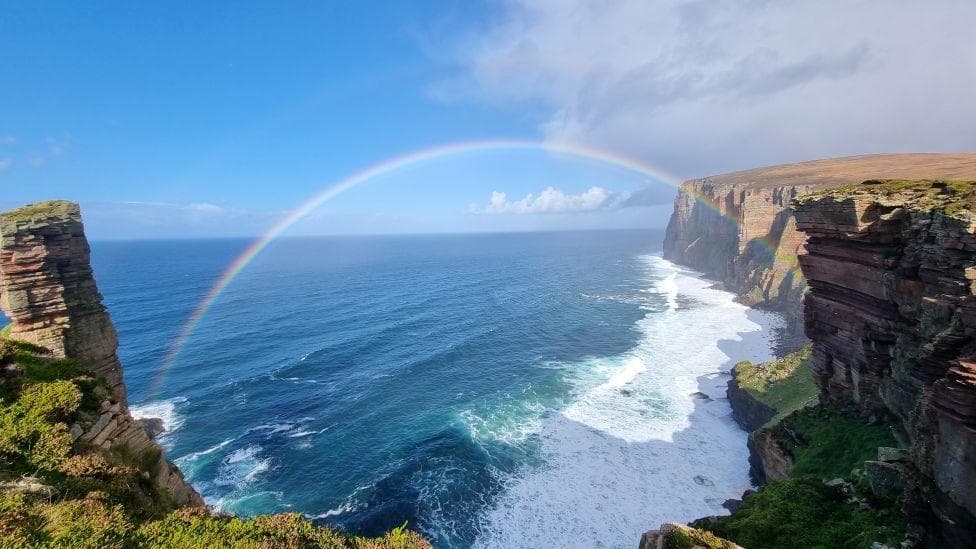 A rainbow between dramatic cliffs over a blue wavy sea and half blue / half cloudy sky.