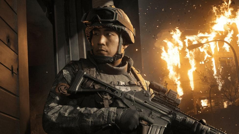 An in-game screenshot showing a male Korean soldier holding a gun with a building on fire behind him.