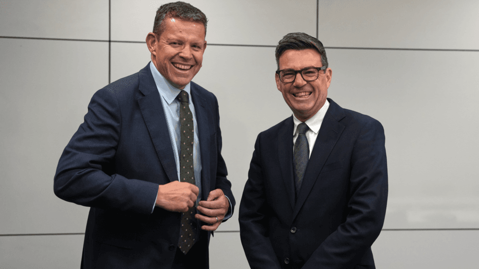 Rhun ap Iorwerth and Andy Burnham smiling. Rhun is on the left and has a dark blue jacket and light blue shirt, with a green tie. Burnham is on the right and has a dark blue jacket, white shirt and green tie. He also has black-framed glasses on.