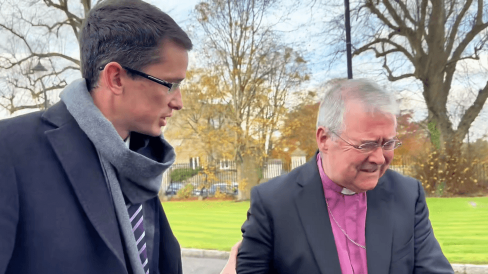 A photo shows Enoch Burke with his hand on the arm of the Church of Ireland Archbishop of Armagh, John McDowell. Burke (left) is wearing a navy suit with a grey scarff and a purple tie. McDowell has a navy suit on with a purple shirt. He has grey hair.