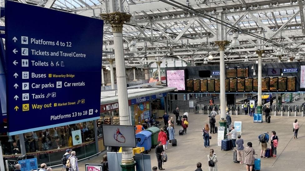 A far shot of the inside of Edinburgh Waverly train station. Boards displaying service times hang over the gates for boarding the platforms. There is a large open area with people milling about holding bags, with shops around the edges of the space. A blue board hangs from the glass ceiling giving directions for toilets, platforms, car parks etc