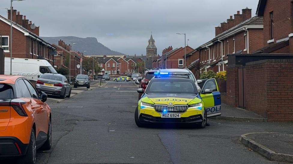 A police car with its door open and blue lights on. It is in a street full of brick houses and a variety of cars. There is a police cordone further done the road with another police car in the distance.
