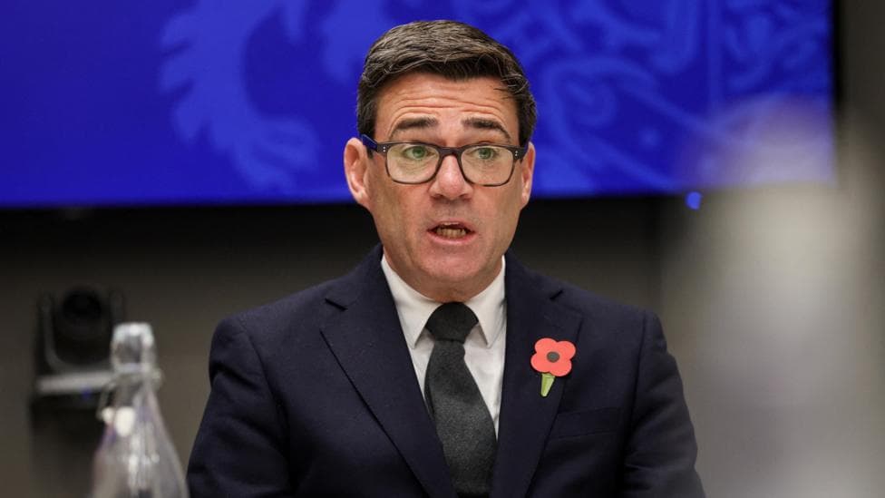 Prime Minister Andy Burnham. He has dark hair and glasses, and is wearing a dark suit, black tie and poppy.