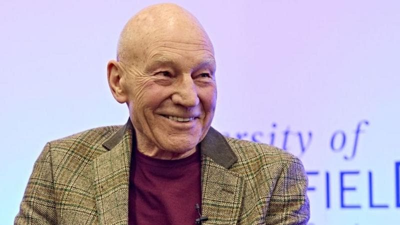 Sir Patrick Stewart sits in a chair smiling. He is wearing a tweed jacket over a dark-coloured t-shirt. Behind him is a partially obscured sign for th University of Huddersfield