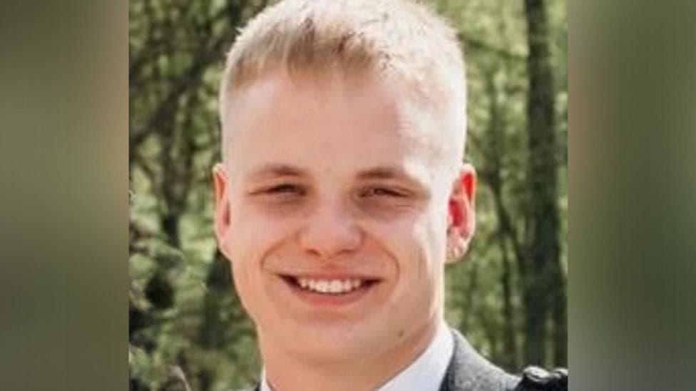 A young man with blonde hair smiles at the camera - he is outside, with trees in the background