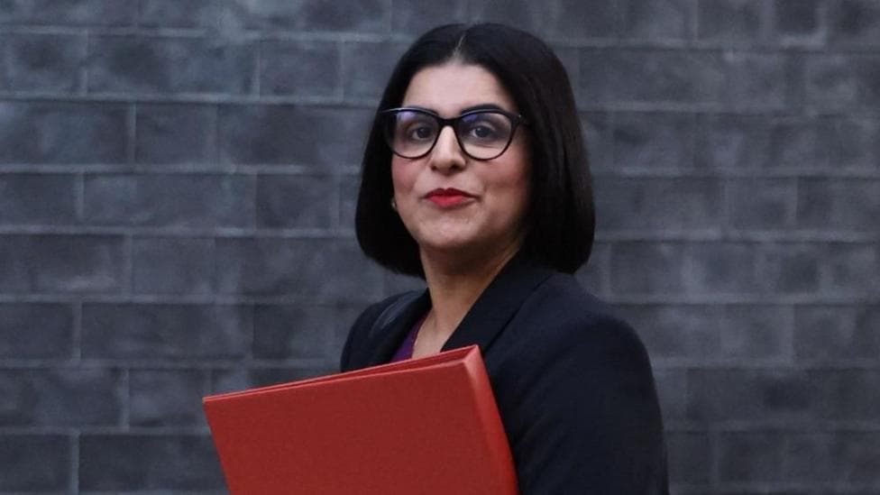 Home Secretary Shabana Mahmood carries a red folder as she walks past a grey brick wall.