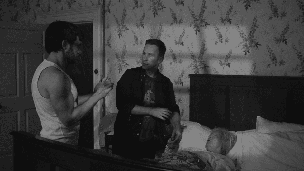 A black and white still from a movie. It depicts a woman with short grey hair lying in a bed. She holds the left hand of a man with short hair wearing a t-shirt and over-shirt. The man look at another man who has short hair and wears a white vest.