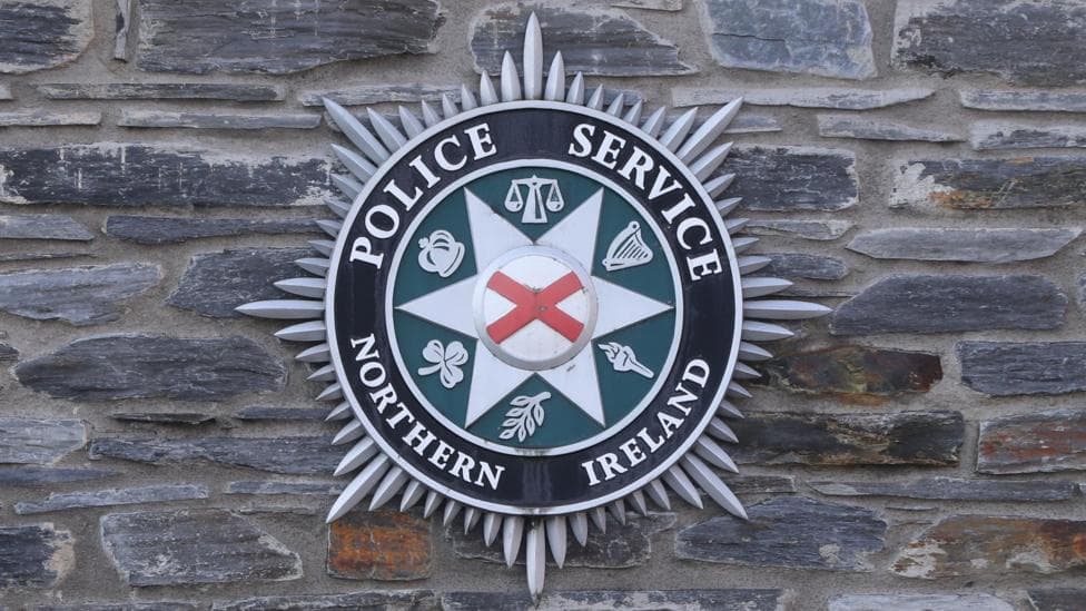 The picture shows the PSNI crest with the words Police Service of Northern Ireland on it. It also has a white star and red diagonal cross, as well as the symbol of the crown, the shamrock, a harp and scales of justioce