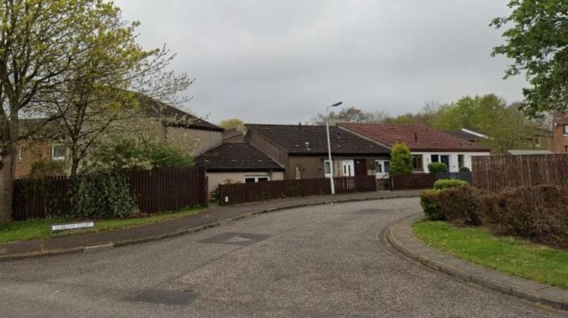 Street view of Dunecht Court. A row of bungalows are behind a fence. The street sign sits on the grass on the corner and there are trees on either side of the road.
