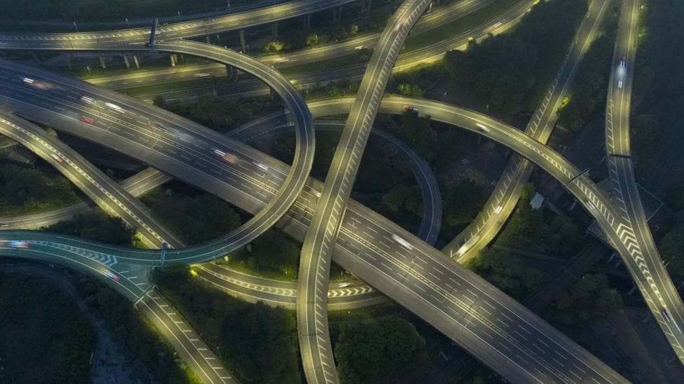 Aerial night-time view of a large motorway interchange, with multiple elevated roads and looping slip roads crossing over one another. Streetlights illuminate the carriageways, creating bright ribbons of light against the dark surrounding landscape. A small number of vehicles are visible as streaks of white and red lights along the roads. Trees and patches of greenery fill the spaces between the sweeping curves of the junction.