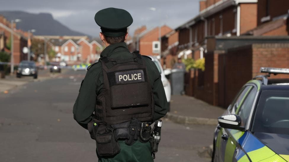 A police officer standing with his back to the camera in a street of red-brick houses. There is a police car beside him.